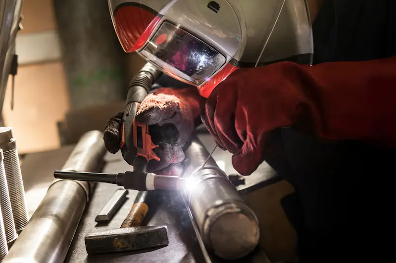 Soudeur portant un masque et des gants rouges effectuant un soudage TIG sur aluminium en atelier.